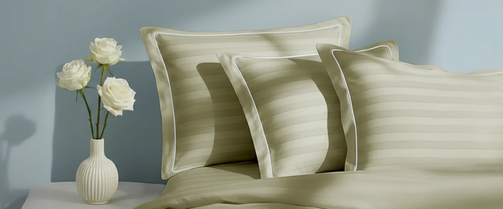 A neatly arranged set of ivory pillow covers from Mulberry Living, with subtle horizontal stripes and white piping detail, placed on a matching bed sheet beside a small white vase holding three white roses against a soft blue background.